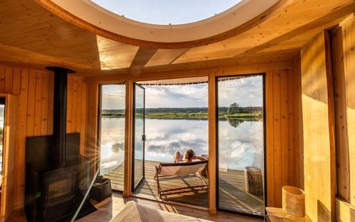 Vue de l’intérieure d‘un chalet en bois lumineux dont les portes s’ouvrent sur une terrasse flottant sur un lac paisible. Une femme est allongée dans un transat sur la terrasse.